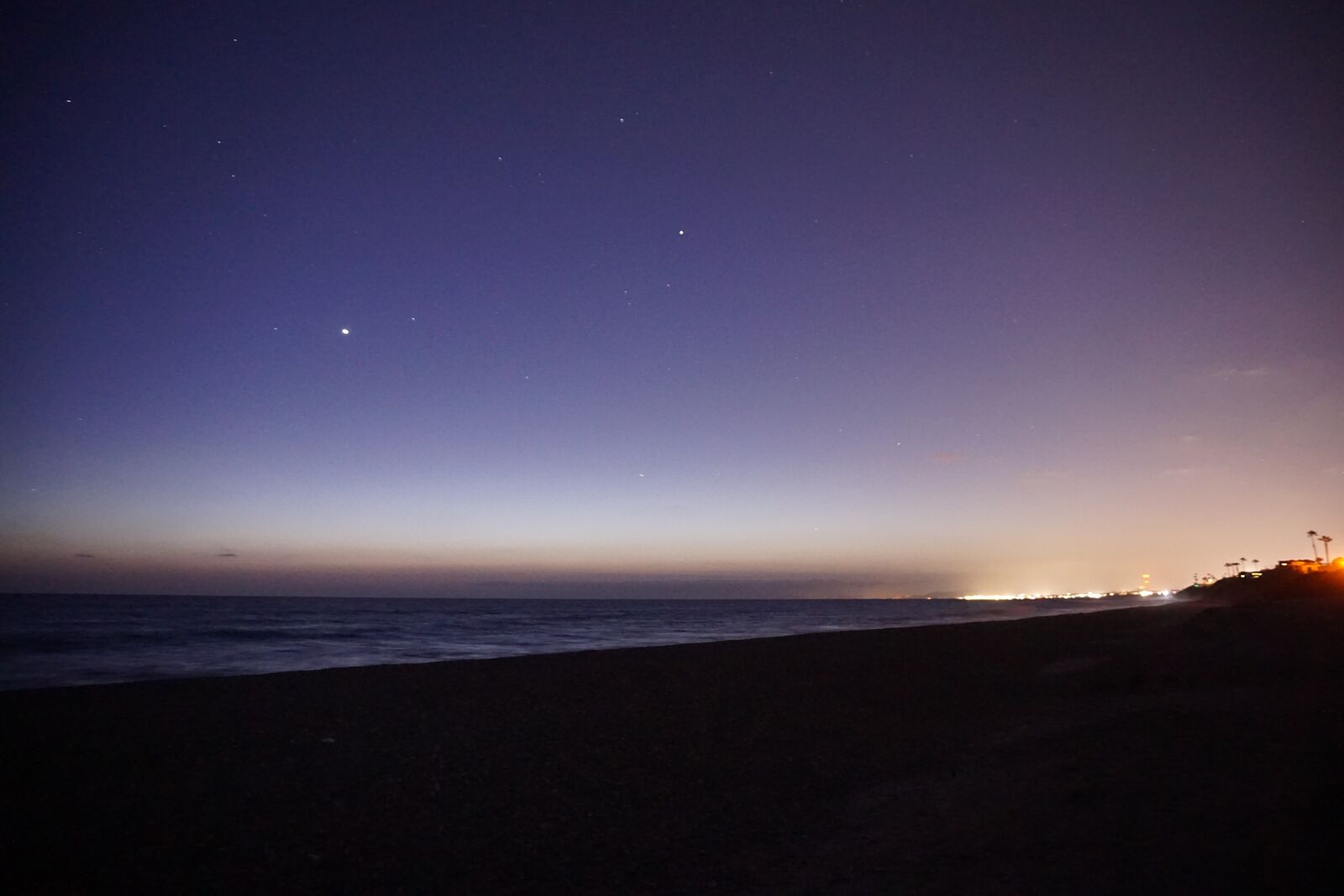 A coastline at blue hour: a deep blue sky fading to orange at the horizon, a dark ocean, and a few lights along the shore.
