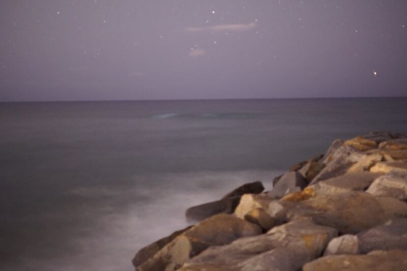 A long exposure of waves smoothed into mist around a jetty of large boulders, under a pale violet night sky.