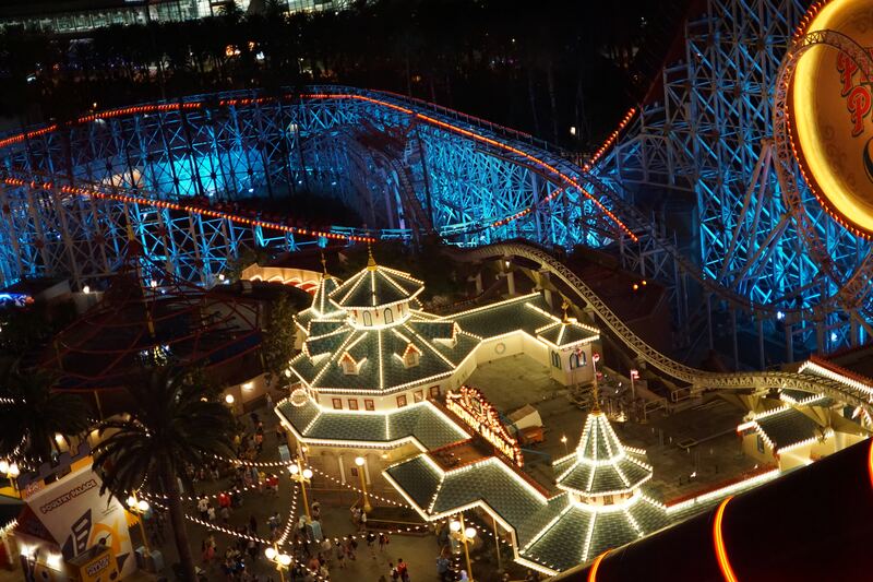 An amusement park at night seen from above: a wooden roller coaster lit blue and a pavilion outlined in strings of white lights.